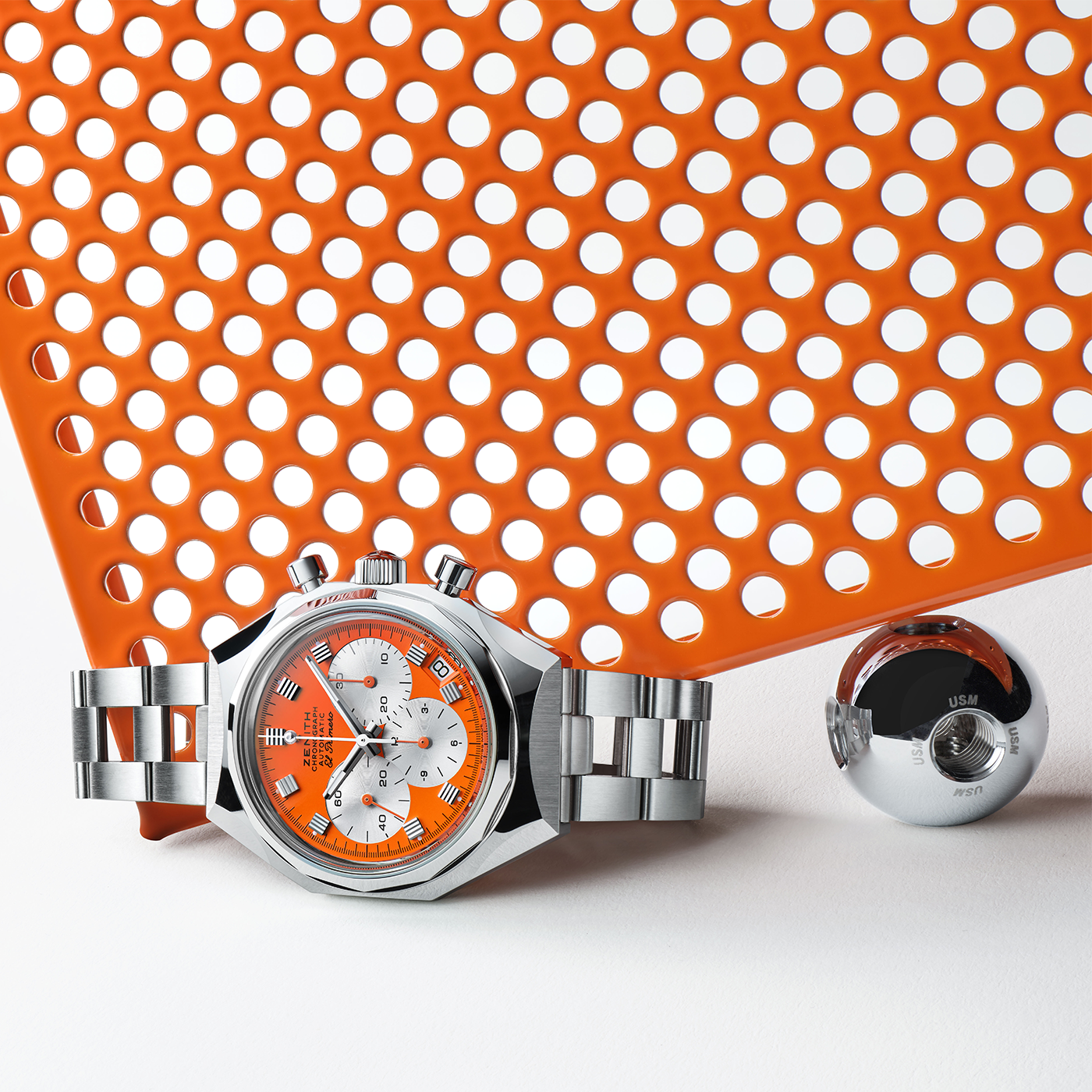 A Zenith Defy Chronograph USM - Pure Orange wristwatch with a silver case and orange-white dial sits by a small round metal object, set on a white surface with an orange perforated backdrop, showcasing Swiss craftsmanship.