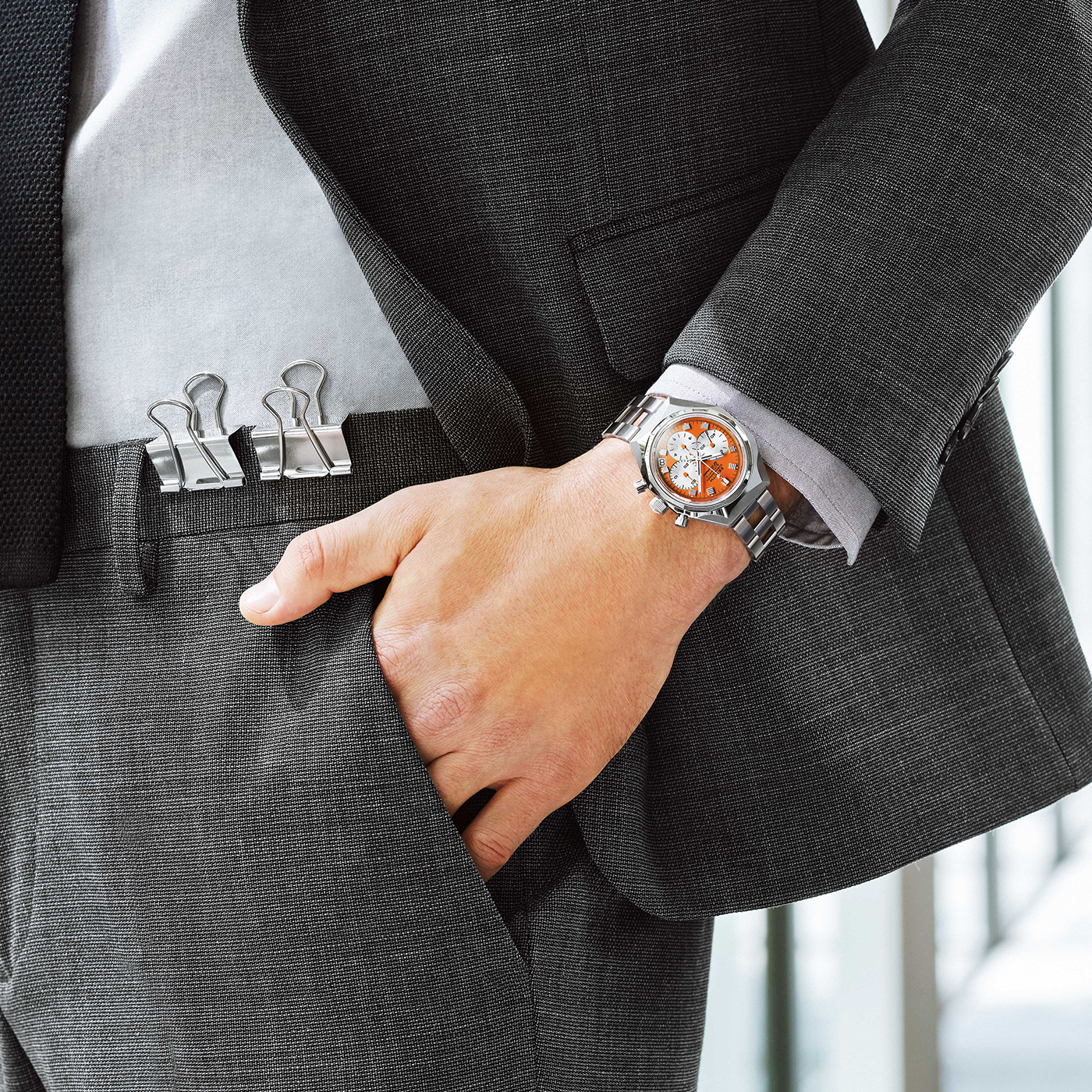 A person in a dark suit with their hand in their pocket, wearing the Zenith Defy Chronograph USM - Pure Orange by Zenith. Three binder clips are attached to the waistband of their suit pants.