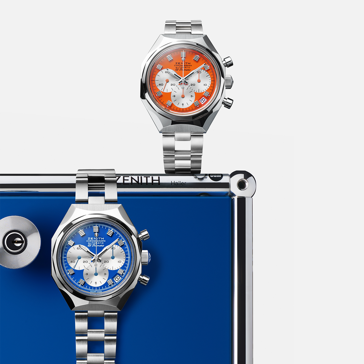 Two Zenith Defy Chronograph USM - Pure Orange wristwatches, one with an orange dial and one with a blue, feature Swiss precision and El Primero 400 movement, displayed on a reflective blue and silver surface.