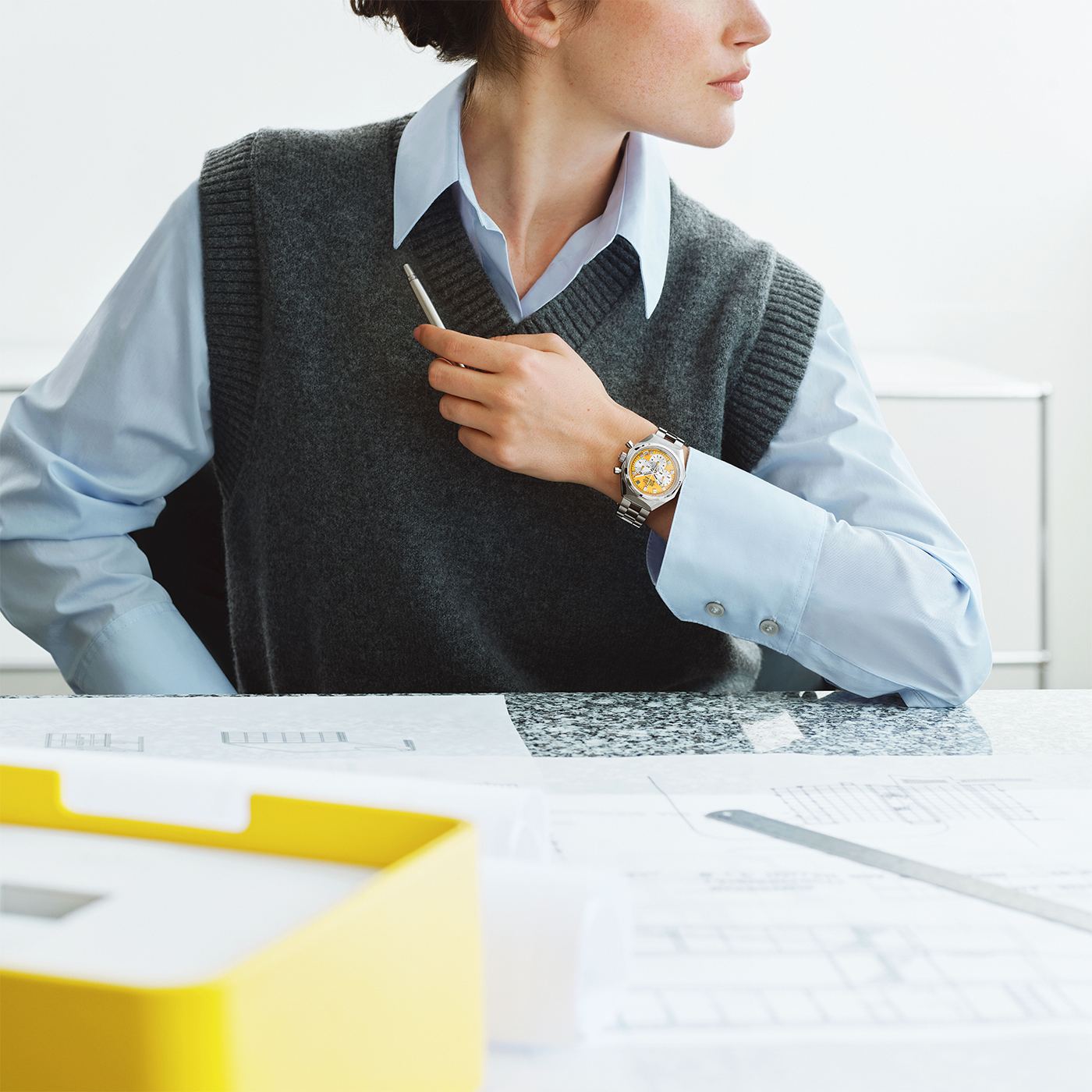 A person in a blue shirt and gray vest, pen in hand at a desk with architectural plans and a yellow file box, mirrors the precision of Swiss mastery embodied by the Zenith Defy Chronograph USM - Golden Yellow.