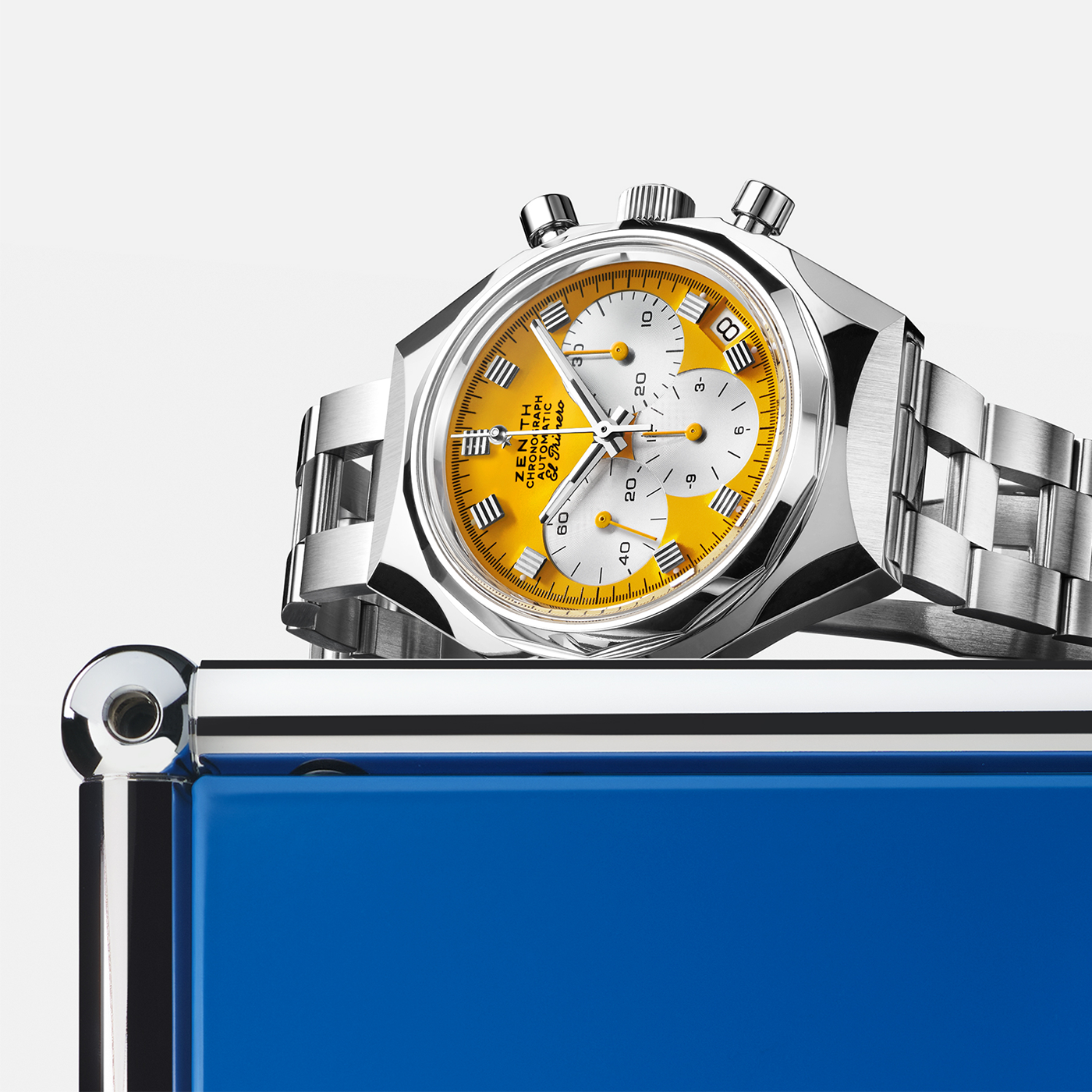 A Zenith Defy Chronograph USM - Golden Yellow by Zenith, featuring a golden yellow and white dial, sits on the edge of a metallic blue rectangular object against a white background.