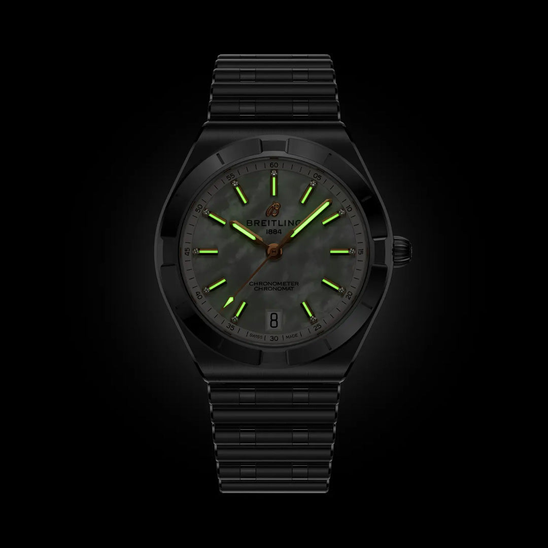 The BREITLING Chronomat Automatic 36, a COSC-certified black sports watch by Breitling, features a dark dial with glowing green markers and hands, metal bracelet, and date at 3 o’clock—shown against a black background.