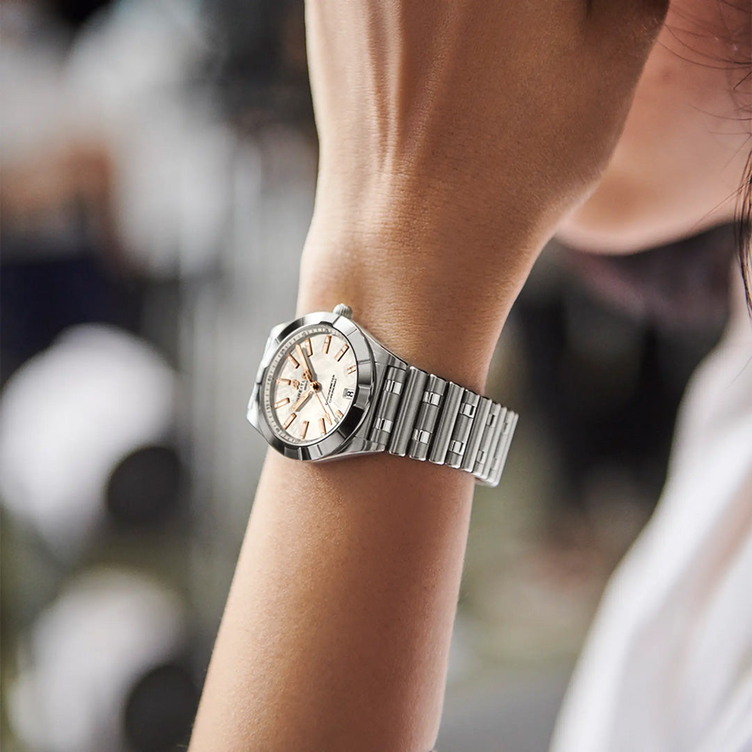 A close-up of a wrist wearing the BREITLING Chronomat 32 by Breitling, featuring a silver analog dial, diamond-set bezel, silver and gold hour markers, and a metal band against a softly blurred background.