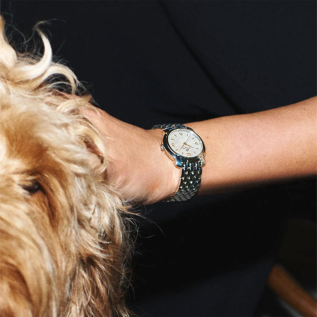 A close-up of a wrist wearing the BREITLING Lady Premier 32 by Breitling, featuring a white face, as the hand rests on a fluffy golden-haired dog. The dark background highlights both the elegant watch and the dog.