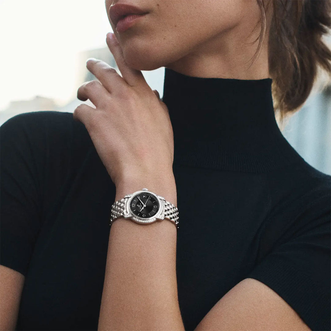 A woman in a black turtleneck touches her neck, showcasing a Breitling Lady Premier 32 with a black dial. Her face is partially visible against a softly blurred background.