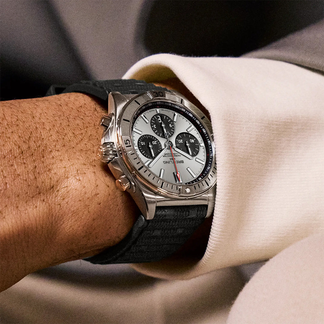 A close-up shows a wrist wearing the stylish BREITLING Chronomat B01 42. This watch features a stainless steel design, in-house Caliber 01, three subdials, and a rotating bezel, with a black strap that complements a white shirt and suit jacket.