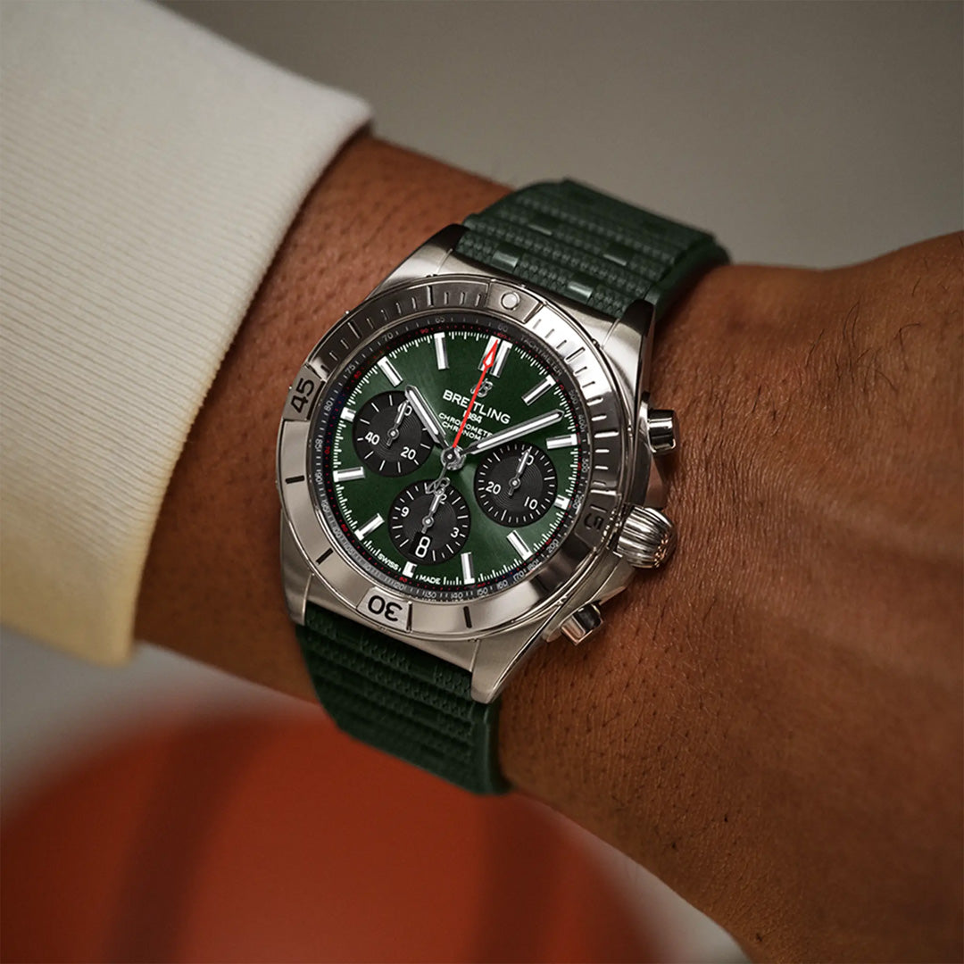 A person is wearing a Breitling Chronomat B01 42 with a silver case and green face and strap. This mechanical chronograph, powered by the Breitling Manufacture Caliber 01, has three sub-dials and a date display. A softly blurred background complements their light-colored long sleeve shirt.