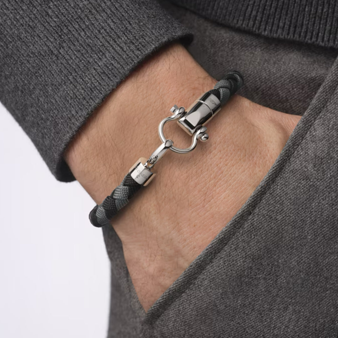 A close-up shows someone wearing an OMEGA Aqua Sailing Nylon Bracelet with a stainless steel clasp. Their hand is in a pocket of a gray sweater, and the bracelet's dark braided pattern adds nautical chic to their look.