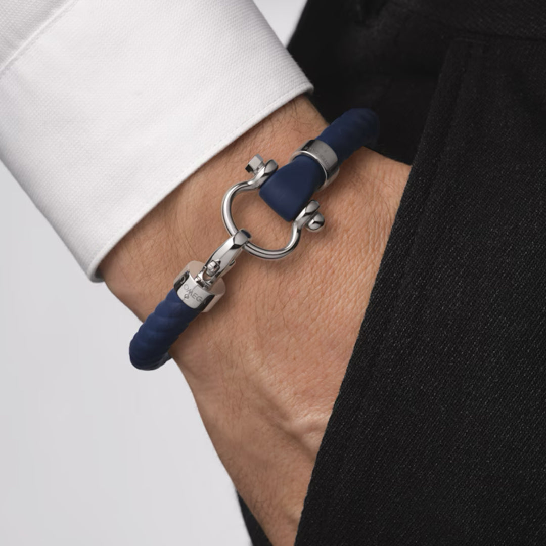 A person in a white shirt and black pants has their hand in their pocket, wearing an OMEGA Aqua Sailing Rubber Bracelet on their wrist, featuring stainless steel accents and a clasp resembling a shackle or horseshoe.