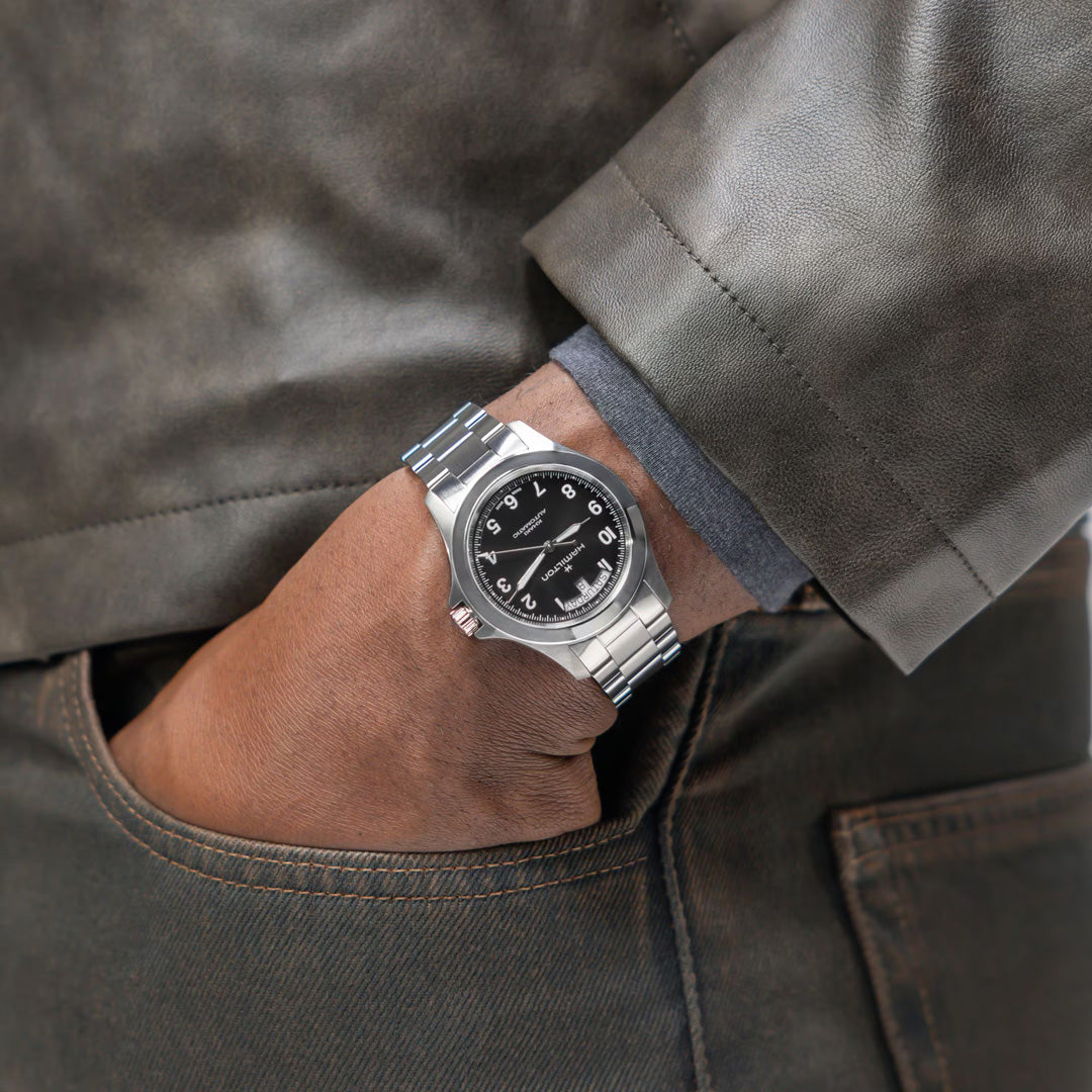 A person wearing a Hamilton Khaki Field King Auto 40mm Watch by Hamilton Watch, featuring a silver case and black dial, has their left hand in brown pants’ pocket, dressed in a gray shirt and dark leather jacket.