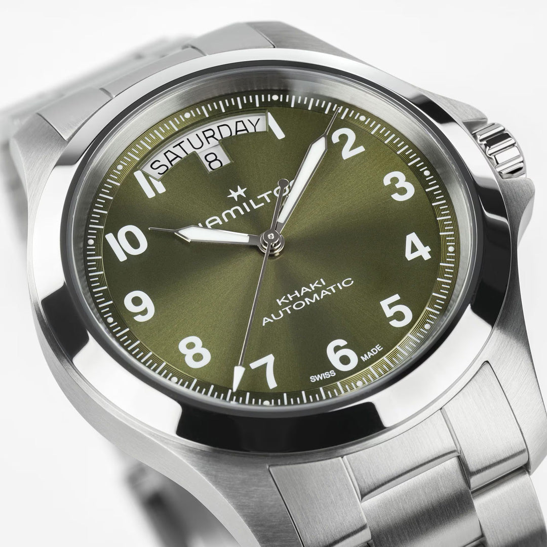 A close-up of the Hamilton Watch Khaki Field King Auto 40mm shows its green dial, silver hour markers and hands, day-date display at "Saturday 8," and stainless steel case with matching bracelet.