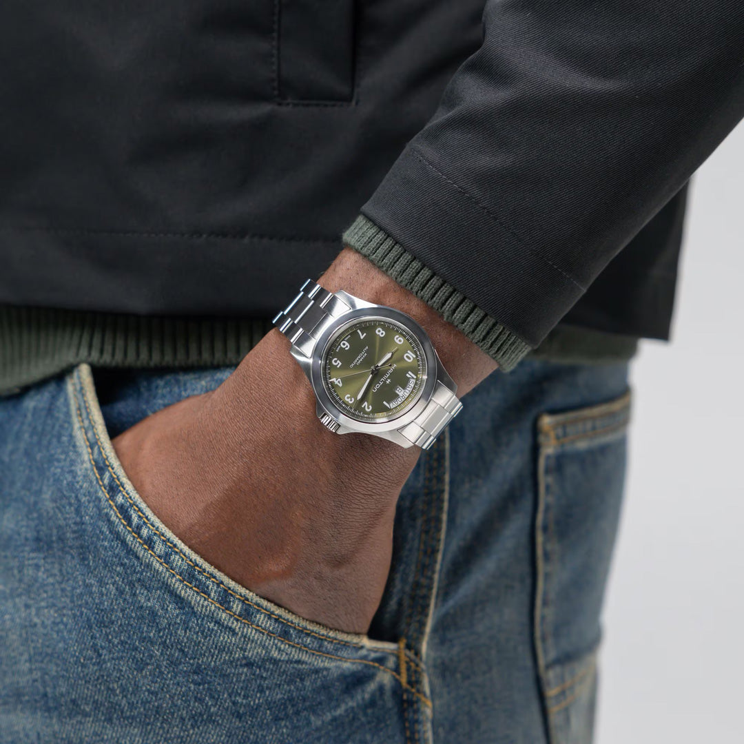 A person in blue jeans, a green sweater, and a black jacket wears the Hamilton Khaki Field King Auto 40mm Watch by Hamilton Watch, featuring a green dial and silver band, with their hand in their pocket.