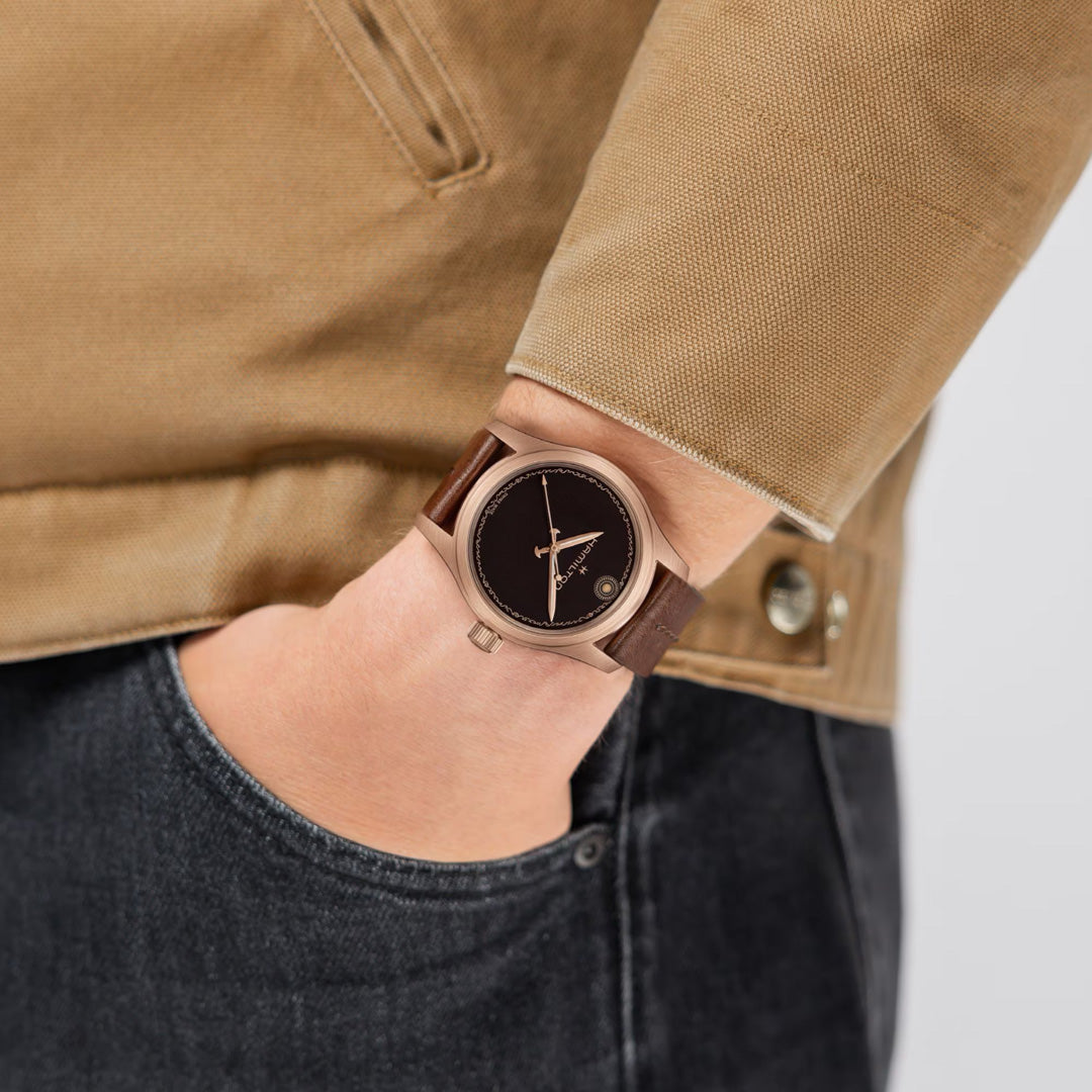 A person wearing a tan jacket and dark jeans has their hand in their pocket, subtly showcasing the Hamilton Khaki Field The Odyssey Limited Edition Auto 42mm Watch—a brown wristwatch with a black face and minimal design by Hamilton Watch, inspired by Christopher Nolan.