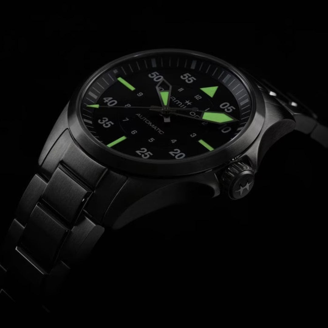 A sleek, dark wristwatch with a metal band is displayed against a black background. The Hamilton Khaki Aviation Pilot Auto 36mm Watch by Hamilton Watch is perfect for aviation enthusiasts, featuring luminescent green hands and markings. Numbers and an automatic movement label decorate the face, while a star embellishes the crown.