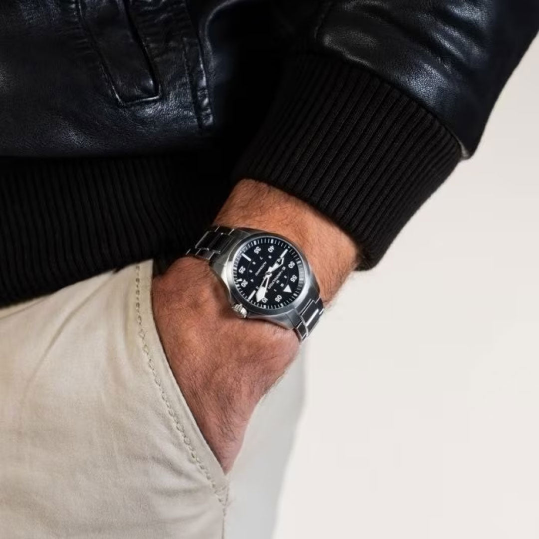 A close-up of a person wearing a black leather jacket and a silver Hamilton Watch wristwatch. The person's hand rests in the pocket of beige pants, showcasing the Hamilton Khaki Aviation Pilot Auto 36mm Watch. This timepiece features automatic movement, a black dial with white hour markers, and enhanced visibility thanks to Super-LumiNova technology.