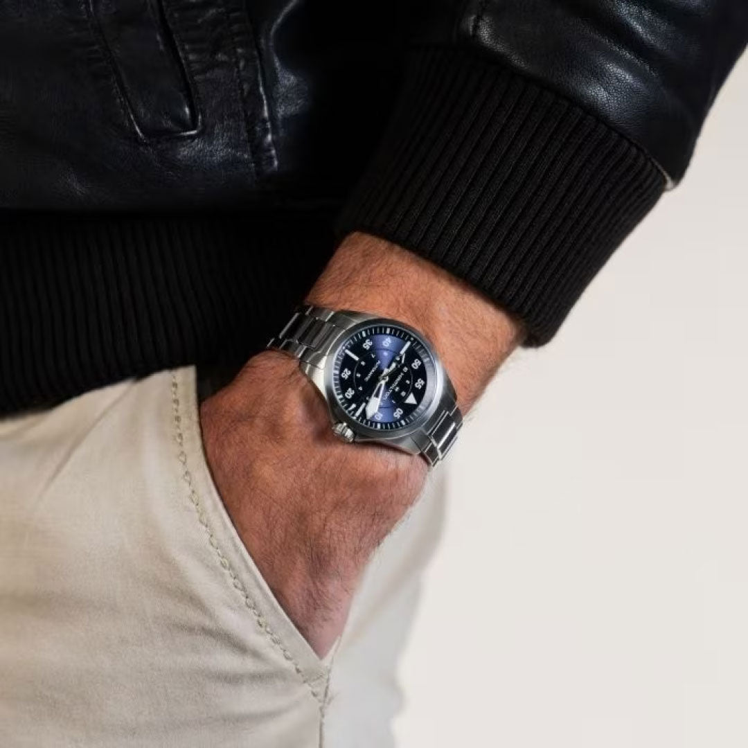 A person wearing the Hamilton Khaki Aviation Pilot Auto 36mm Watch, which has a stainless steel band with a black dial and silver markings, boasting an automatic movement, partially tucks their hand into the pocket of light-colored pants. They are also donning a black leather jacket.