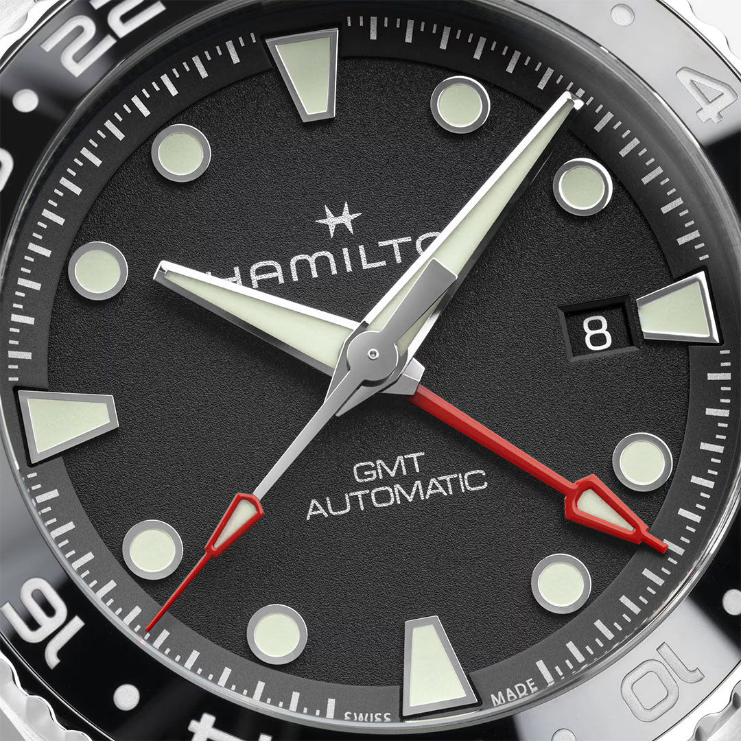 Close-up of the Hamilton Khaki Navy Scuba Auto GMT 43mm Watch by Hamilton Watch shows luminous markers, bold hands, a red GMT hand, and date window at 3 o'clock. The black bezel with white numerals emphasizes its diving-inspired design.