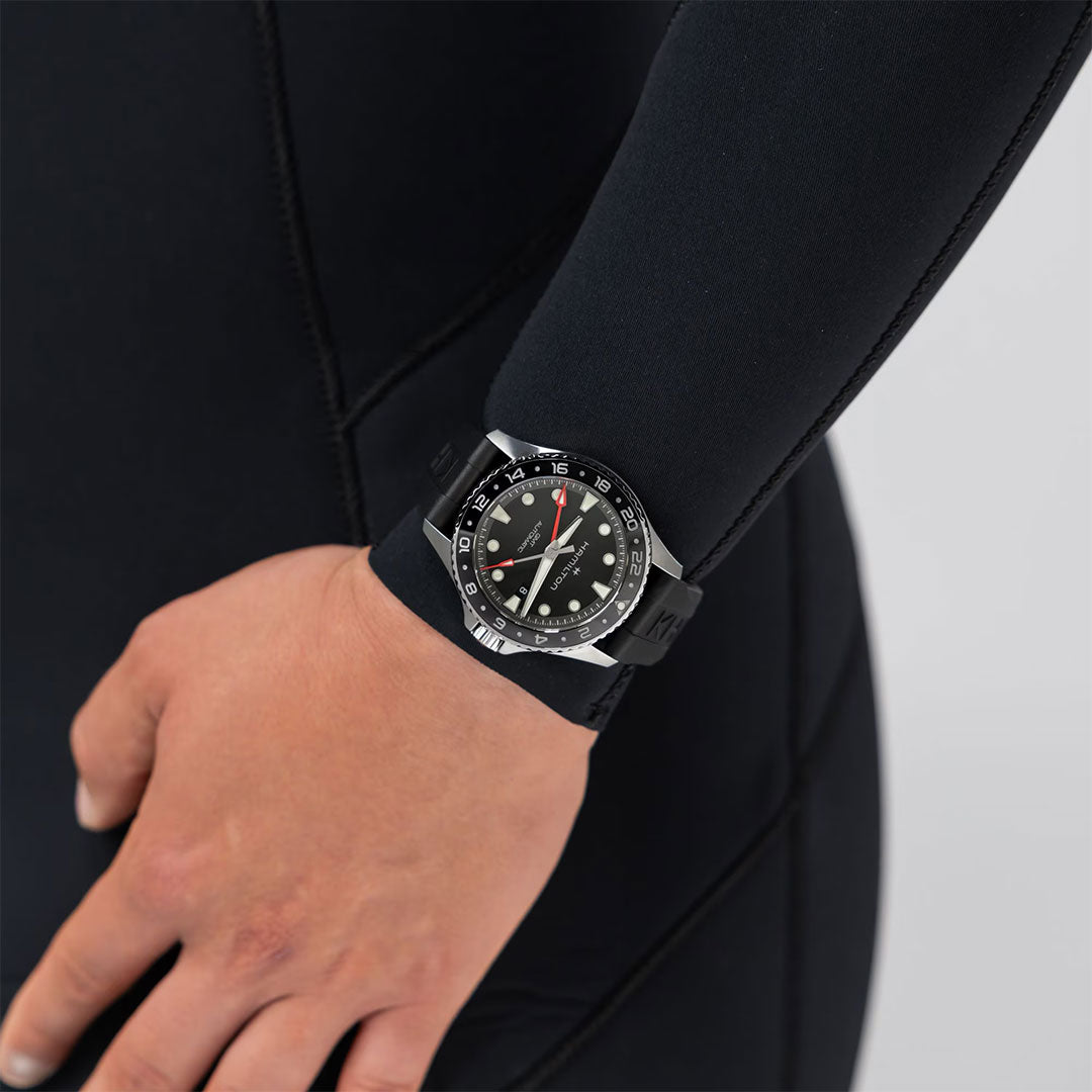 A person in a black wetsuit is shown from the side, their left arm bent, wearing a Hamilton Khaki Navy Scuba Auto GMT 43mm Watch by Hamilton Watch with a black dial on their wrist.