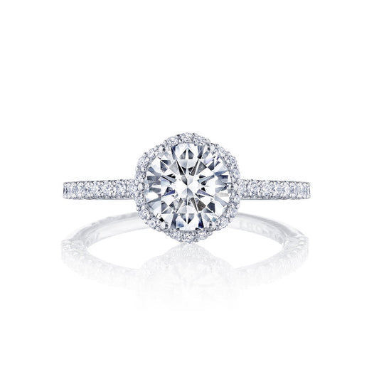 This Tacori Petite Crescent 18K White Gold Round Brilliant Semi Mount with Diamond Band and Halo Engagement Ring showcases a central round diamond surrounded by a halo of smaller diamonds. The band, adorned with diamonds, enhances its shine against a white background, making this piece truly exquisite.