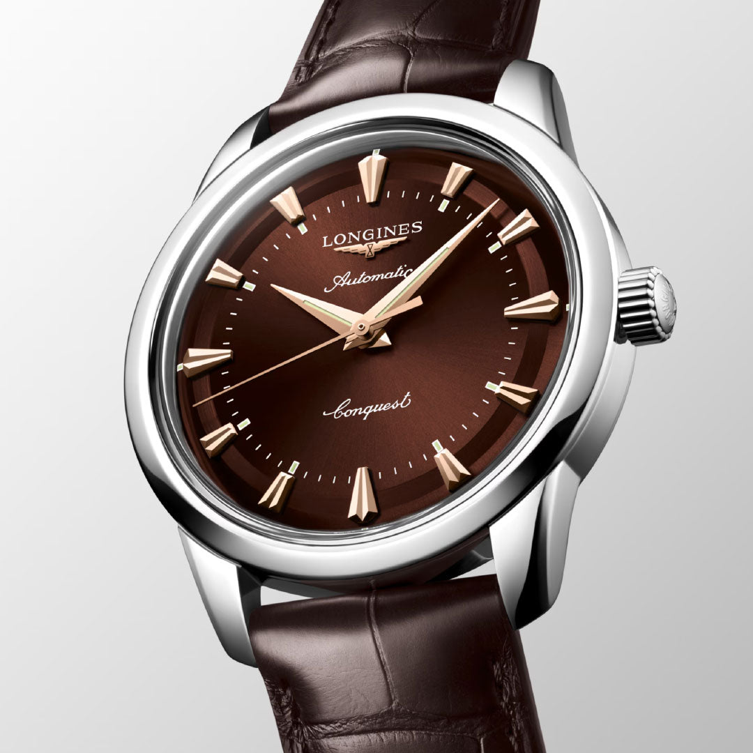 The Longines Conquest Heritage Automatic 40mm Watch features a brown dial with gold markers, a silver case, and a brown leather strap, all showcased against a light backdrop for vintage appeal.