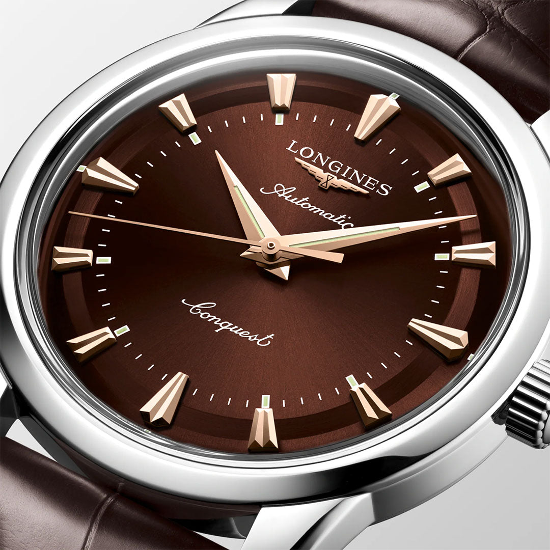 A close-up of the Longines Conquest Heritage Automatic 40mm Watch featuring a brown dial, gold hour markers and hands, a brown leather strap, silver casing, and visible Longines branding for a vintage look.