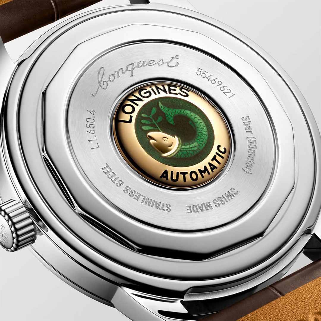 A close-up of the Longines Conquest Heritage Automatic 40mm Watch shows its stainless steel back with an engraved fish and green wave logo on a gold center, inscribed with “Swiss Made” and design details echoing Longines’ vintage style.