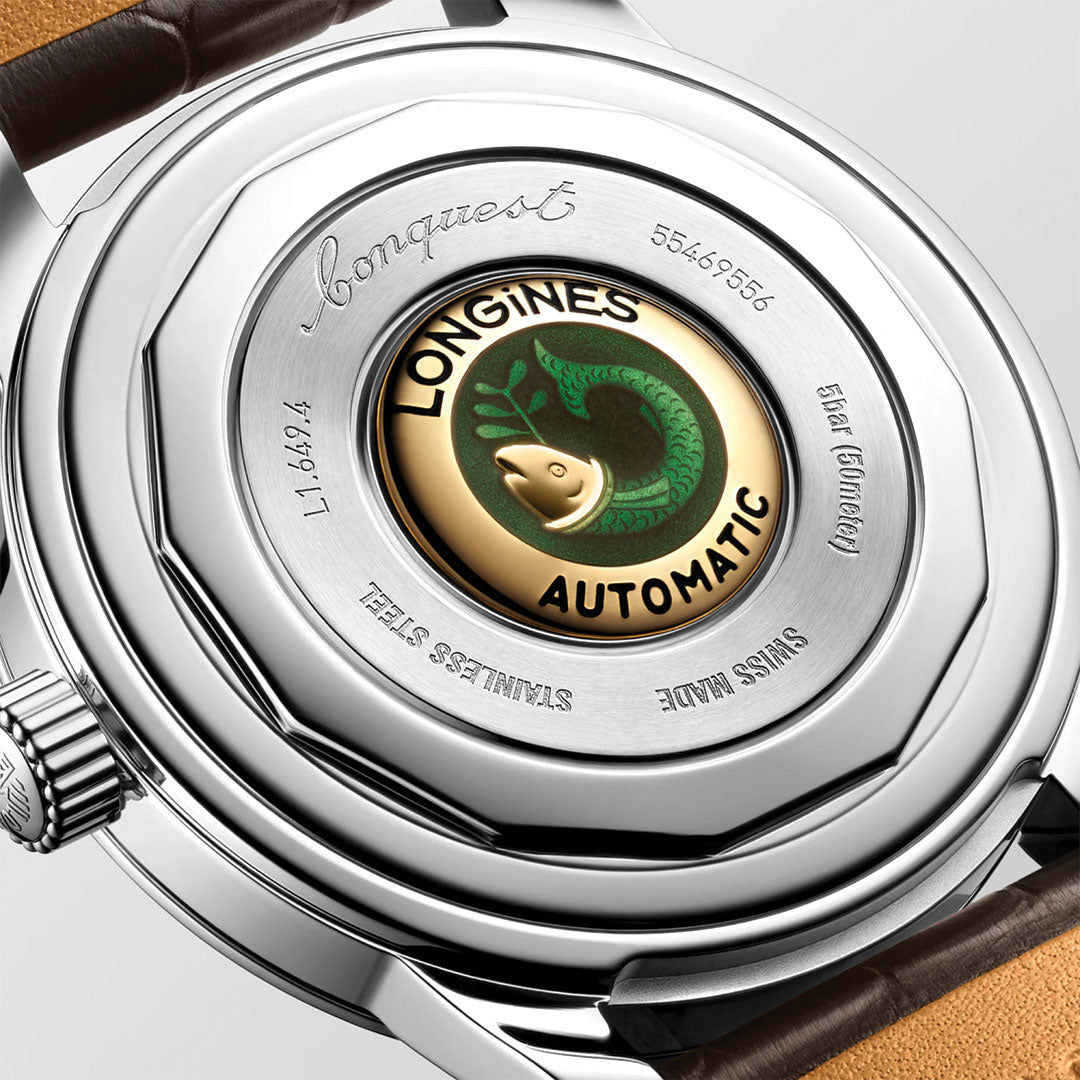 Close-up of the Longines Conquest Heritage Automatic 38mm Watch by Longines, highlighting its silver case with engraved text, gold center medallion with a green fish and “Longines Automatic,” plus a vintage-style brown leather strap.
