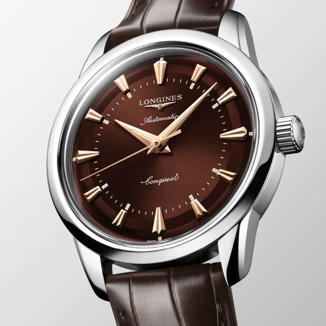 A close-up of the Longines Conquest Heritage Automatic 38mm Watch, featuring a brown dial, gold hands and hour markers, brown leather strap, and a polished silver case with crown on the right.