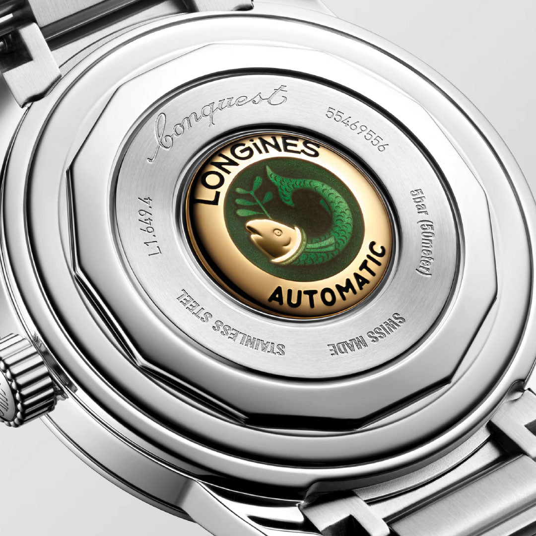 Close-up of the Longines Conquest Heritage Automatic 38mm Watch, showing its stainless steel case, engraved details, and a gold and green emblem with a fish and "LONGINES AUTOMATIC," highlighting its vintage design.
