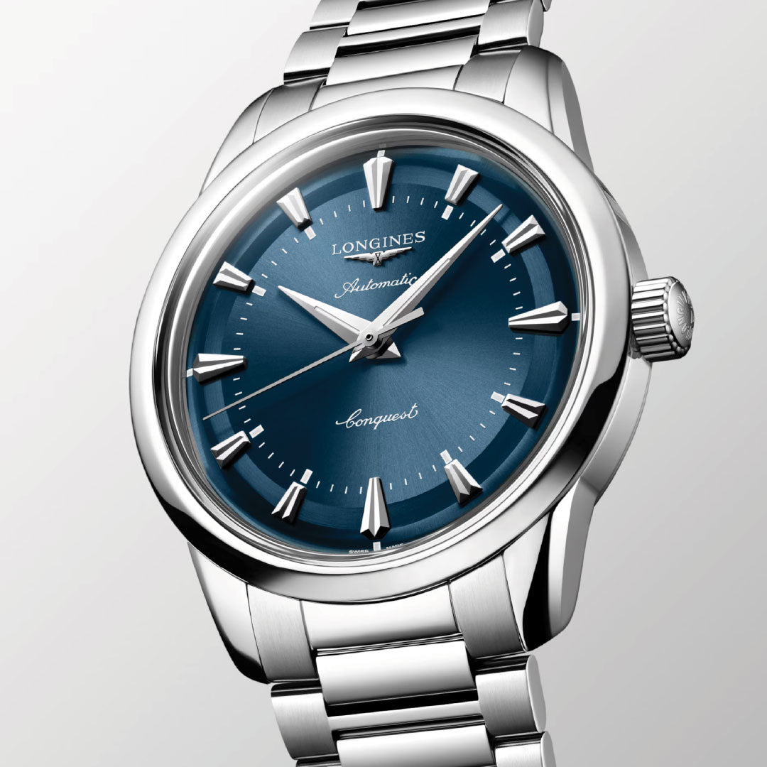 A close-up of the Longines Conquest Heritage Automatic 38mm Watch in stainless steel, featuring a blue dial, silver hour markers, and metal bracelet, with "Longines," "Automatic," and "Conquest" on the face.
