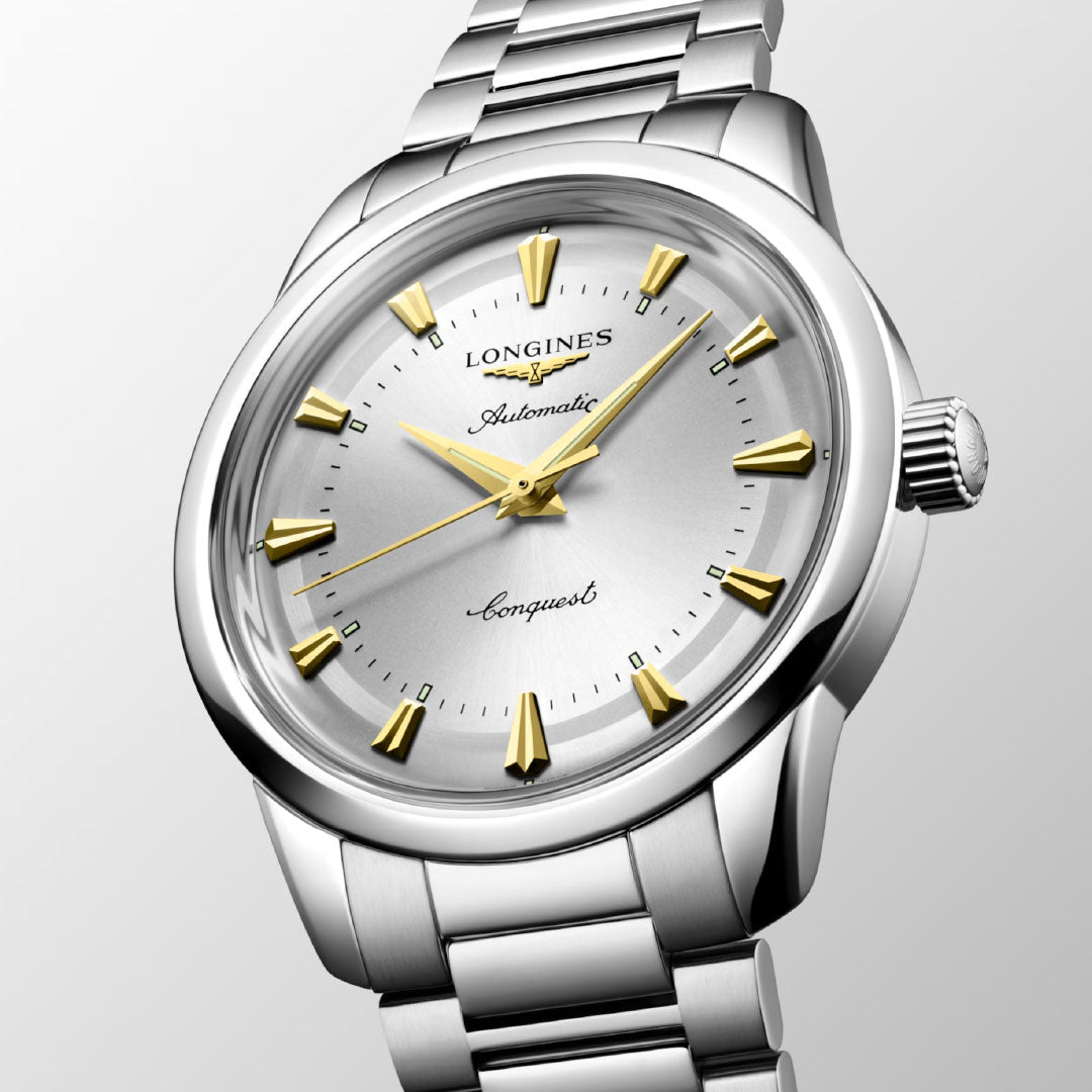 The Longines Conquest Heritage Automatic 40mm Watch features a silver dial, gold-tone hour markers and hands, and a stainless steel bracelet. Shown angled on a light background, its vintage design is elegantly highlighted.