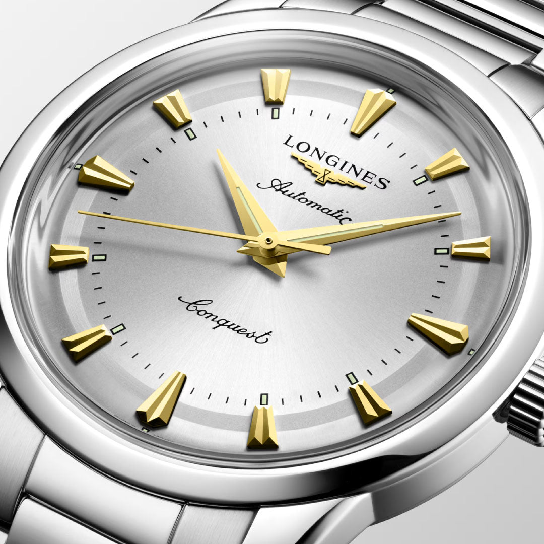 Close-up of the Longines Conquest Heritage Automatic 40mm Watch, featuring gold-tone hands and markers, vintage details, a silver dial, stainless steel bracelet, and "Longines Automatic Conquest" on the face.