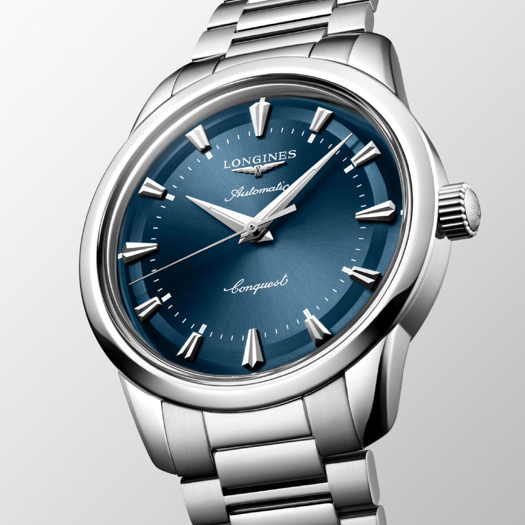 A close-up of the Longines Conquest Heritage Automatic 40mm Watch features a blue dial, silver hour markers, and a stainless steel band, highlighting Longines' modern watchmaking against a light gradient background.