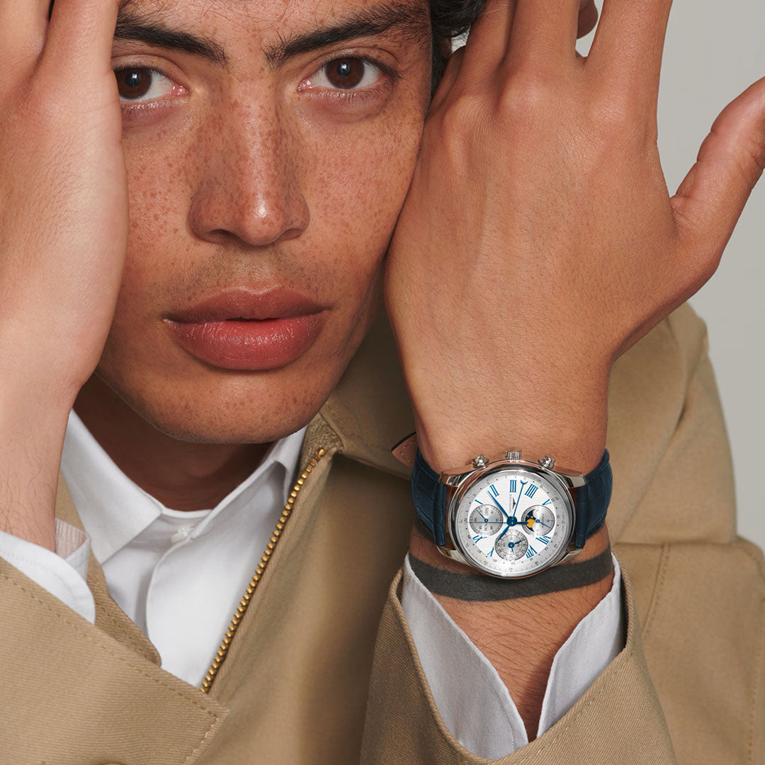 A person with freckles poses with one hand on their face, wearing a beige jacket, white shirt, and the Longines Master Collection Automatic Moonphase 40mm Watch—a refined nod to Longines watchmaking craftsmanship.