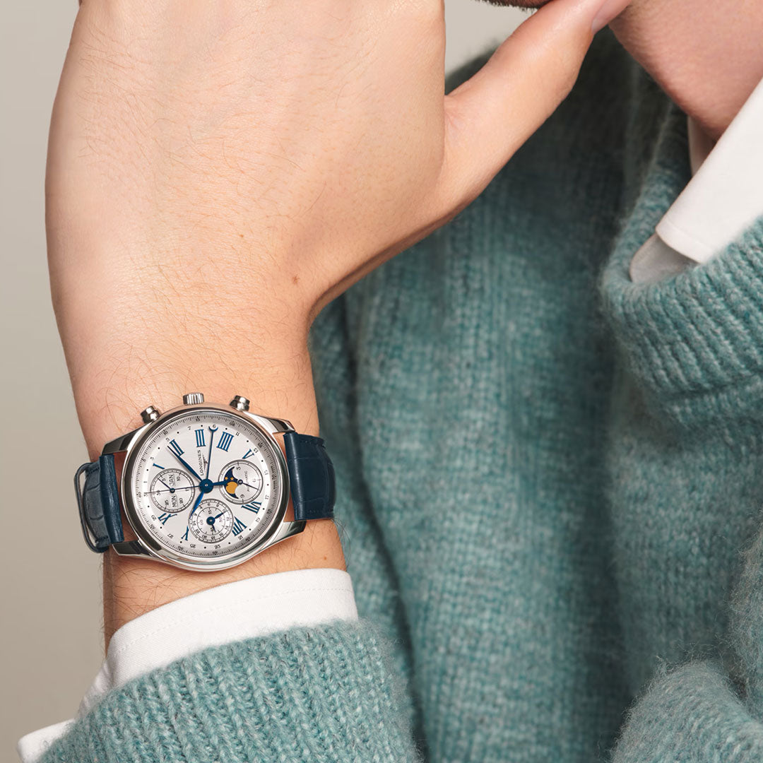A person in a teal sweater and white shirt displays their wrist wearing the Longines Master Collection Automatic Moonphase 40mm Watch, featuring a silver chronograph, white dial, blue hands, Roman numerals, and a dark blue leather strap.