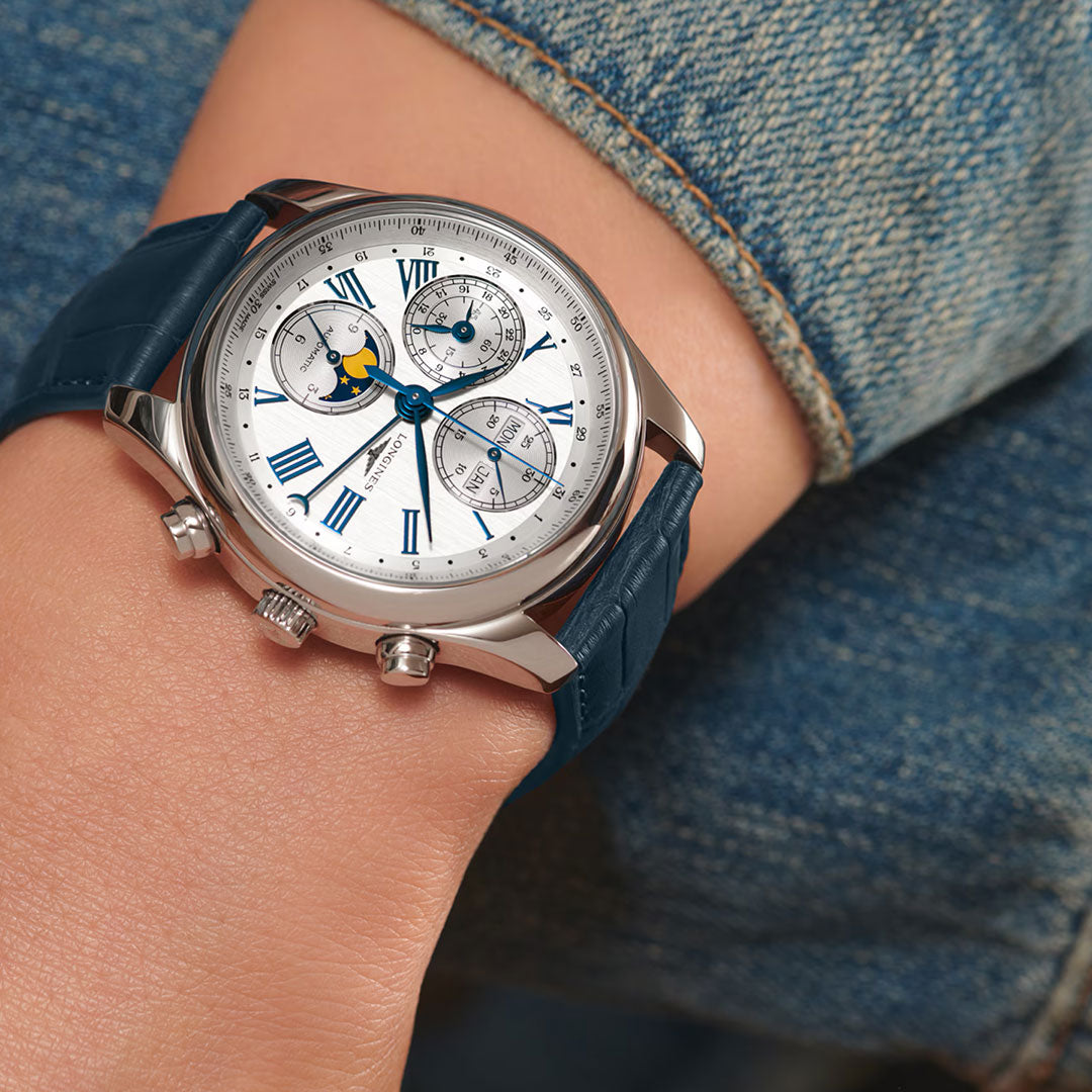 A close-up of a wrist wearing the Longines Master Collection Automatic Moonphase 40mm watch with a white dial, blue Roman numerals, moonphase display, and blue leather strap. The wearer is dressed in blue denim, emphasizing Longines craftsmanship.