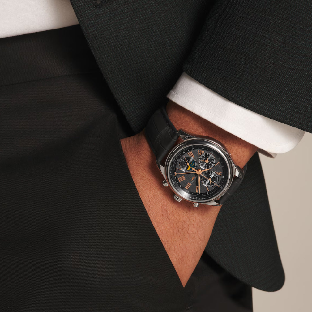 A person in a dark suit with a white cuff and hand in pocket, wearing a Longines Master Collection Chrono Moonphase Automatic 42mm watch with a dark strap, featuring an intricate dial design showcasing impeccable watchmaking craftsmanship.