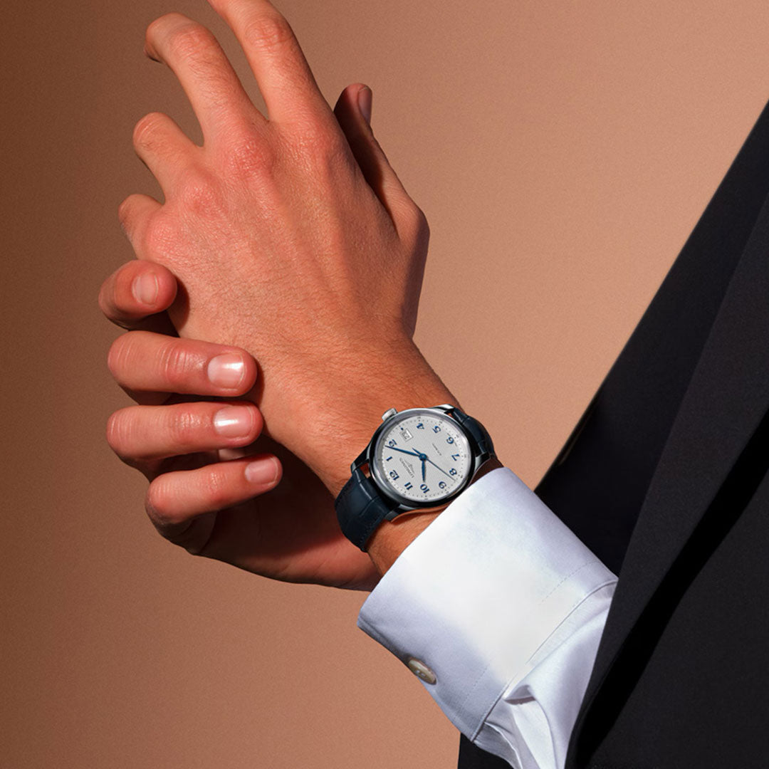 A person in a dark suit and white shirt adjusts their left wrist, showcasing the Longines Master Collection Automatic 39mm Watch with a blue leather strap, set against a neutral brown background.