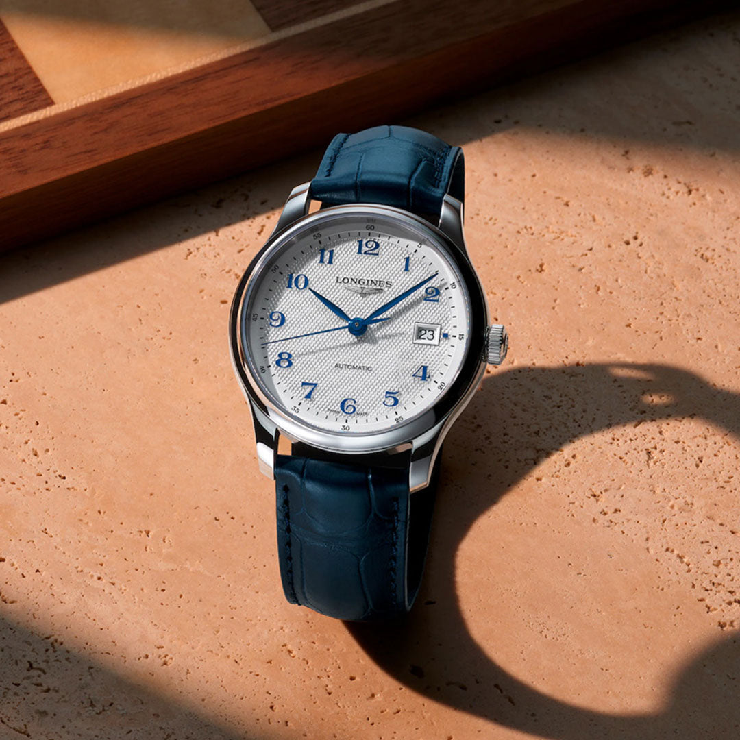 The Longines Master Collection Automatic 39mm Watch features blue hour markers and hands, a white textured dial, and a blue leather strap, showcased on a beige surface beside wood, reflecting Longines' horological craftsmanship.