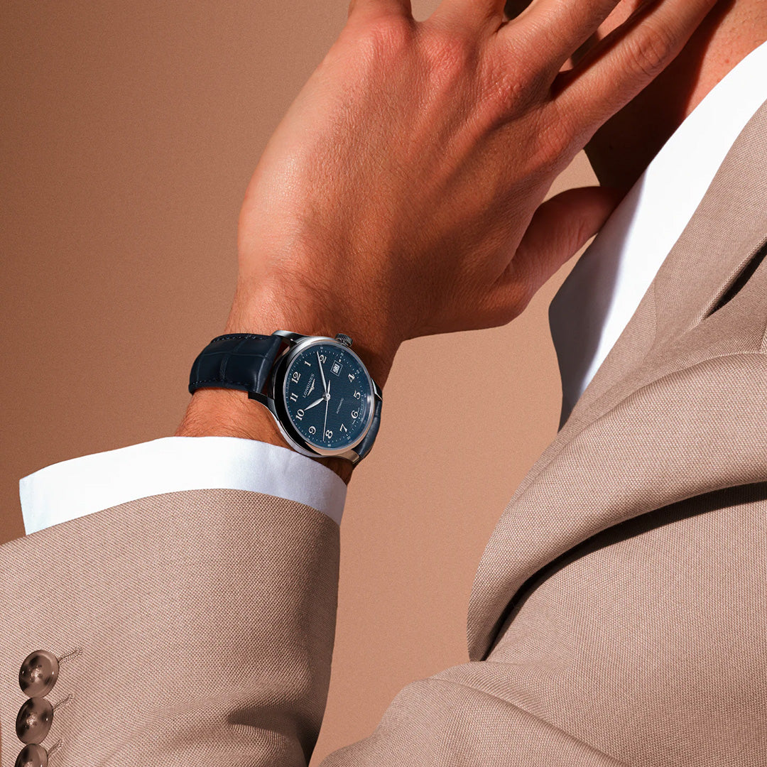 A person dressed in a beige suit and white shirt showcases the Longines Master Collection Automatic 39mm Watch by Longines, featuring a black leather strap and silver casing, exemplifying exceptional horological craftsmanship as they touch their face against a neutral background.