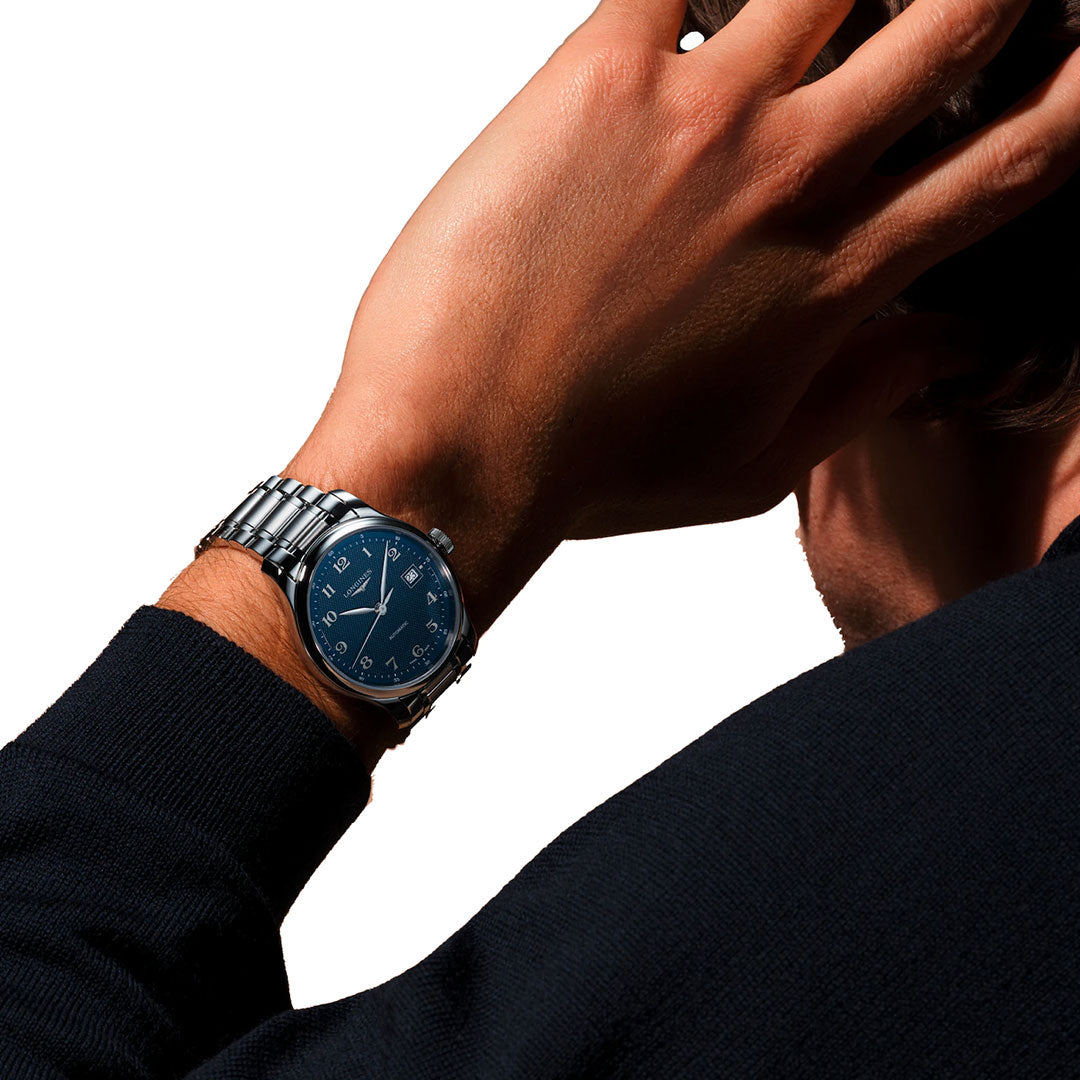 A person wearing a dark sweater displays their wrist, showcasing the Longines Master Collection Automatic 39mm Watch. The silver timepiece features a striking blue face, elegantly highlighted against a plain white background—emphasizing the renowned artistry of Longines Swiss watchmaking and the timeless allure of luxury watches.