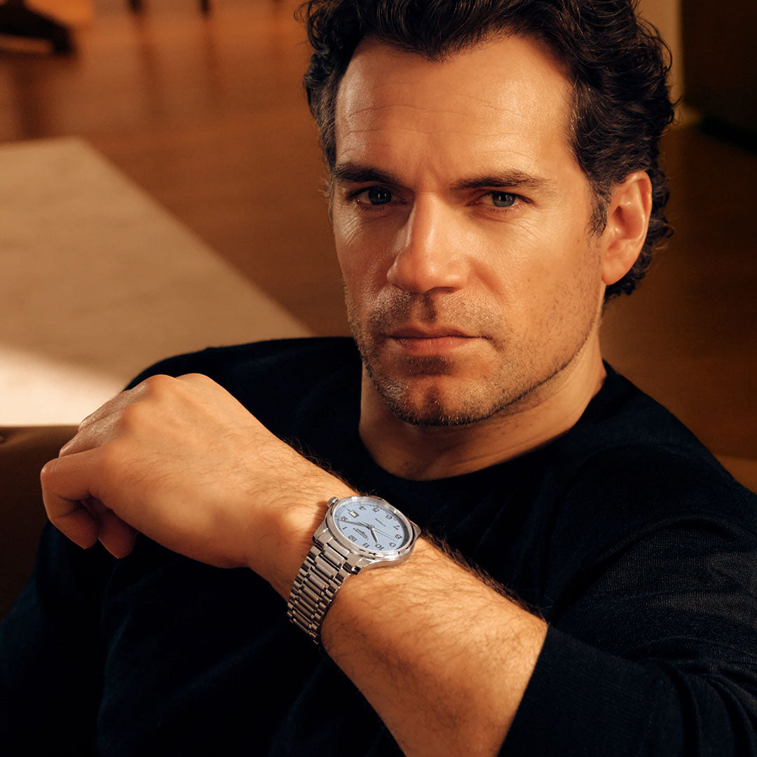 A man with short curly hair, dressed in a black shirt, sits on a chair and looks at the camera, showcasing a silver Longines Master Collection Automatic 41mm Watch on his left wrist—an elegant creation by Longines that exemplifies the artistry of Swiss watchmaking.
