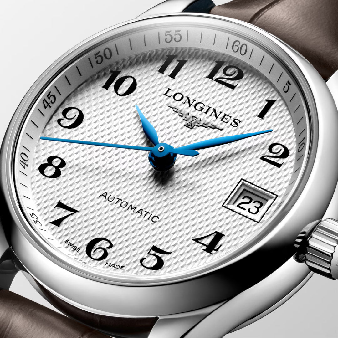 Close-up of the Longines Master Collection Automatic 25.5mm Watch by Longines, featuring a textured white dial, blue hands, black Arabic numerals, date window at 3 o’clock, and a brown leather strap—a luxury timepiece.