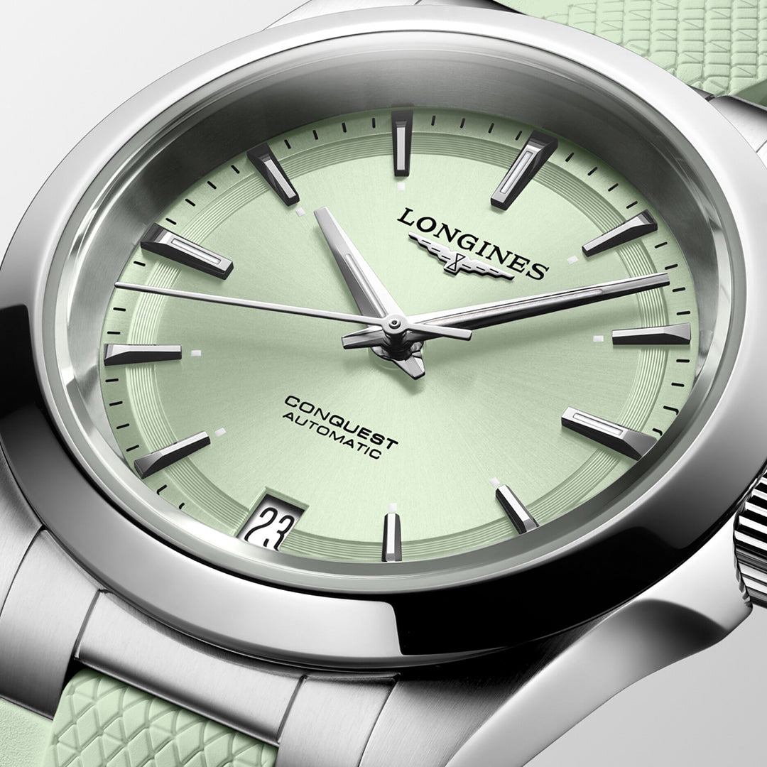 A close-up of the Longines Conquest Automatic 34mm Watch highlights its horological excellence with a mint green dial. The watch features silver hour markers, hands, and a date display at the 3 o'clock position. Its strap is textured in a complementary mint green color.