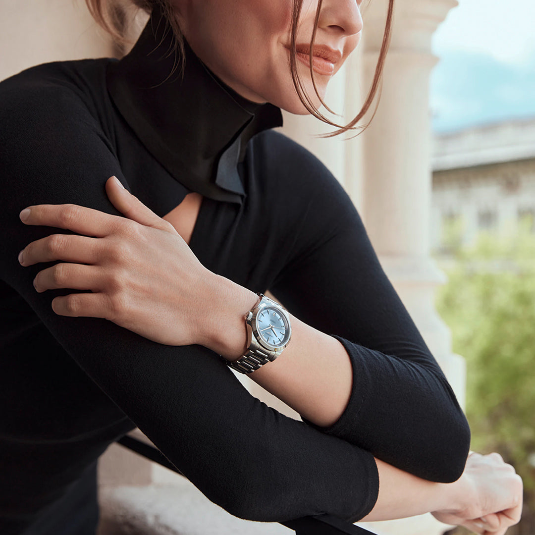 A person wearing a sleek black top leans against a stone railing, arms crossed. Adorning their wrist is a Longines Conquest Automatic 34mm Watch, featuring a silver finish with a blue face. Their hair falls casually across their face, set against the backdrop of a blurred outdoor scene.