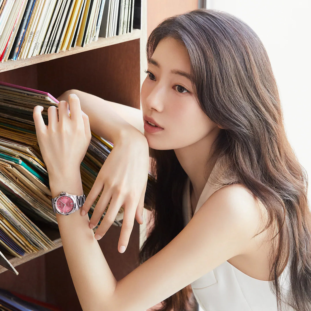 A woman with long hair, wearing a sleeveless top and a Longines Conquest Automatic 34mm watch with a pink face, leans against a shelf filled with records. She gazes thoughtfully into the distance, embodying horological excellence.