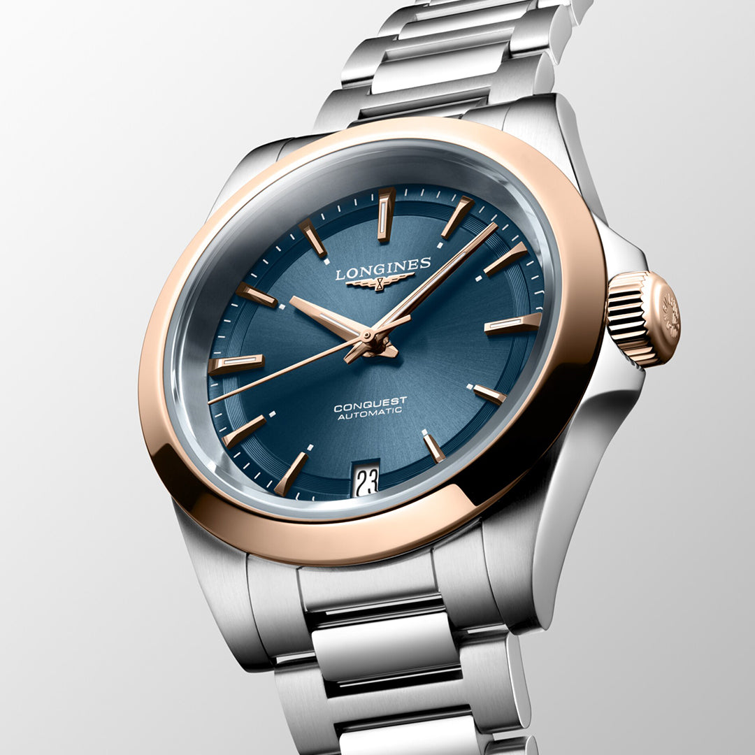 The Longines Conquest Automatic 34mm Watch is a sophisticated timepiece showcasing horological excellence. It features a metallic band, blue dial, and gold bezel, complemented by gold hour markers and hands. The brand "LONGINES" is elegantly displayed along with "Conquest Automatic," and there's a date display at the bottom of the dial.