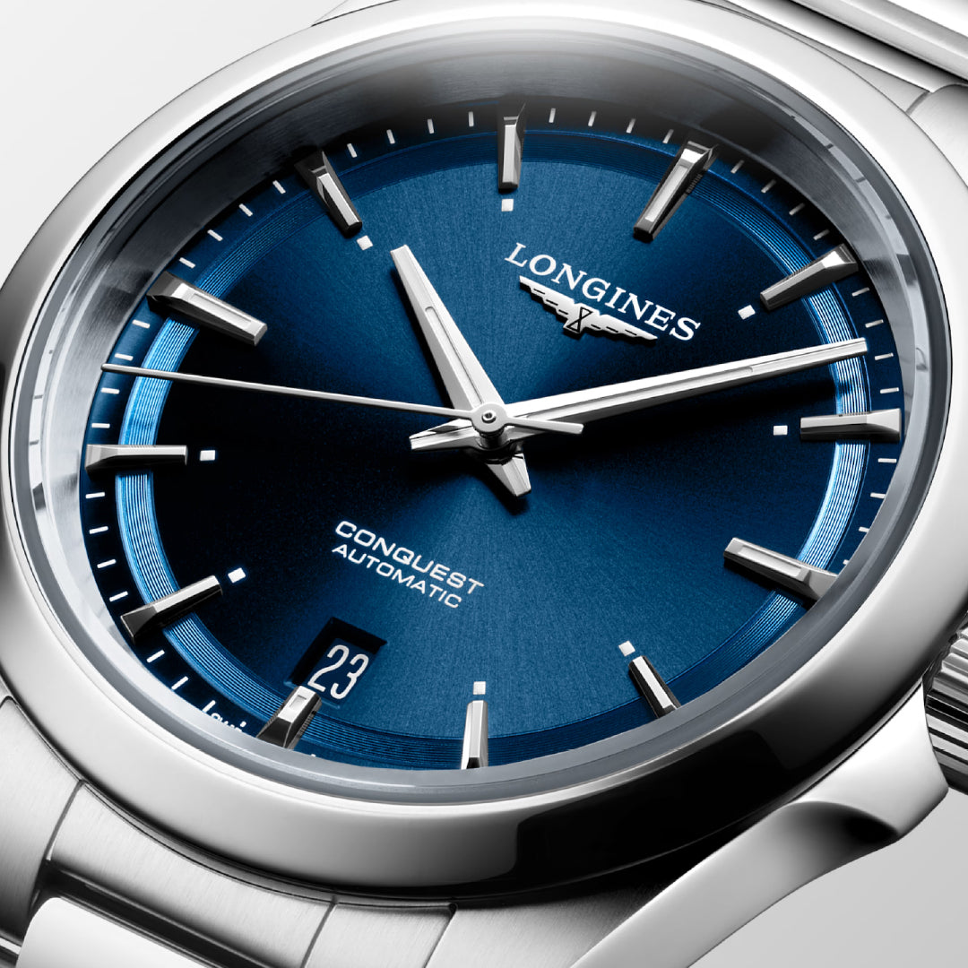Close-up of the Longines Conquest Automatic 38mm Watch featuring a stainless steel bracelet, blue dial with silver markers and hands, date at 6 o'clock, scratch-resistant sapphire crystal, and Longines logo below 12.