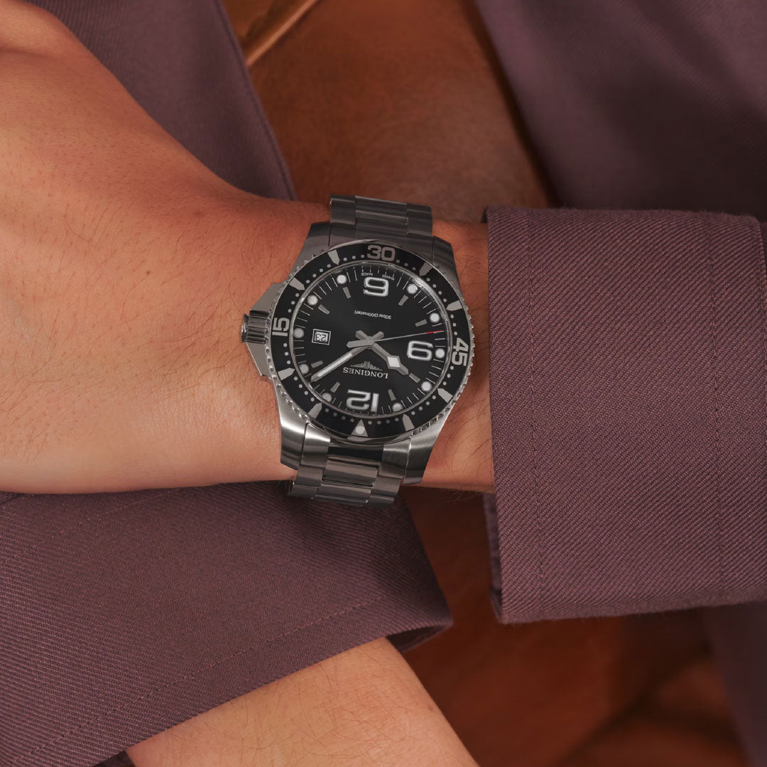 A person in a long-sleeve burgundy shirt wears a Longines Hydroconquest Automatic 41mm watch with a silver metal band, black dial, white numerals, and unidirectional rotating bezel, resting their arm across their lap.