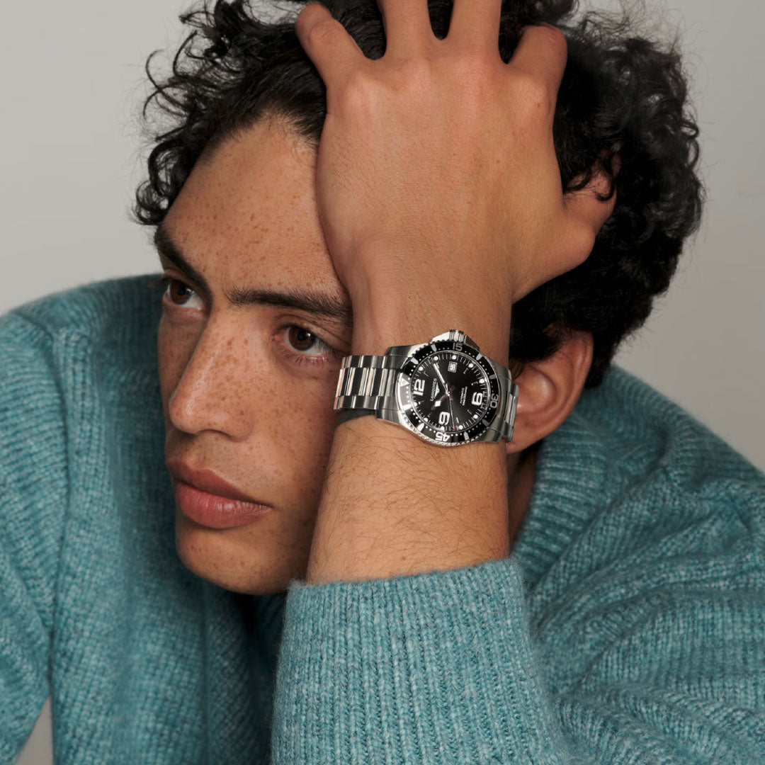 A person with curly hair and freckles, in a teal sweater, rests their head on their hand, showing off the Longines Hydroconquest Automatic 41mm Watch by Longines with bold markings and a unidirectional rotating bezel.